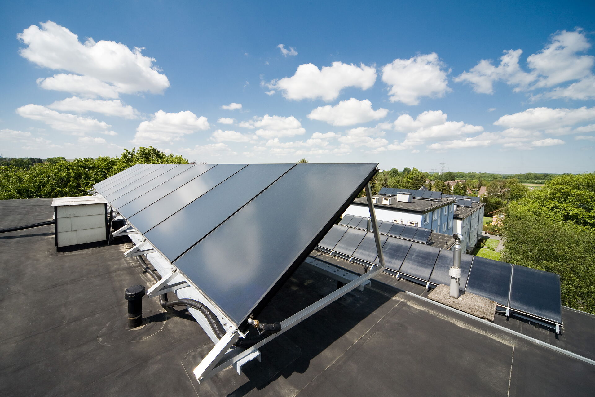 Mehrere Solarmodule auf dem Flachdach eines Gebäudes, ausgerichtet zur Sonne mit Blick über eine grüne Wohngegend und blauen Himmel mit Wolken.