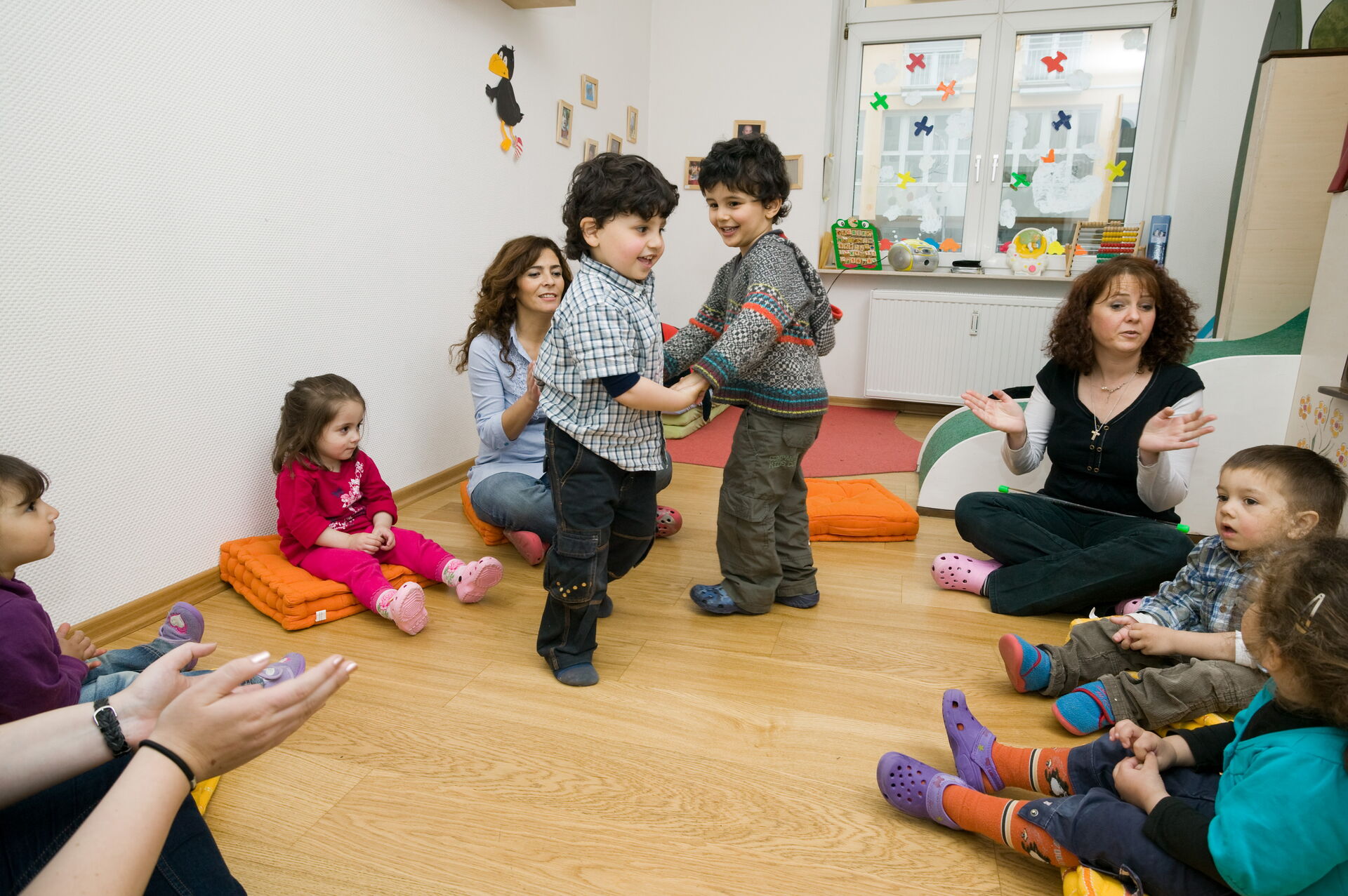 Mehrere Kinder spielen und tanzen gemeinsam im Kreis in einem Gruppenraum, während zwei Betreuerinnen die Aktivität begleiten und andere Kinder im Kreis sitzen.