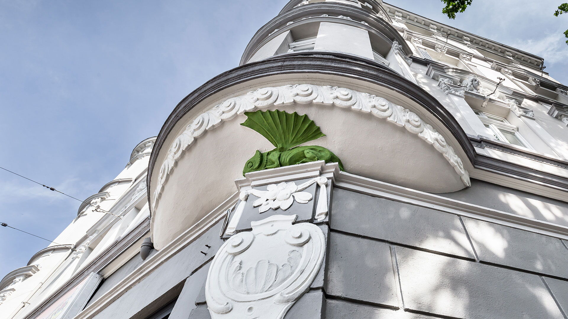 Detailaufnahme einer historischen Gebäudeecke mit weißer Stuckfassade und grünem dekorativem Ornament oberhalb eines Eingangsbereichs.