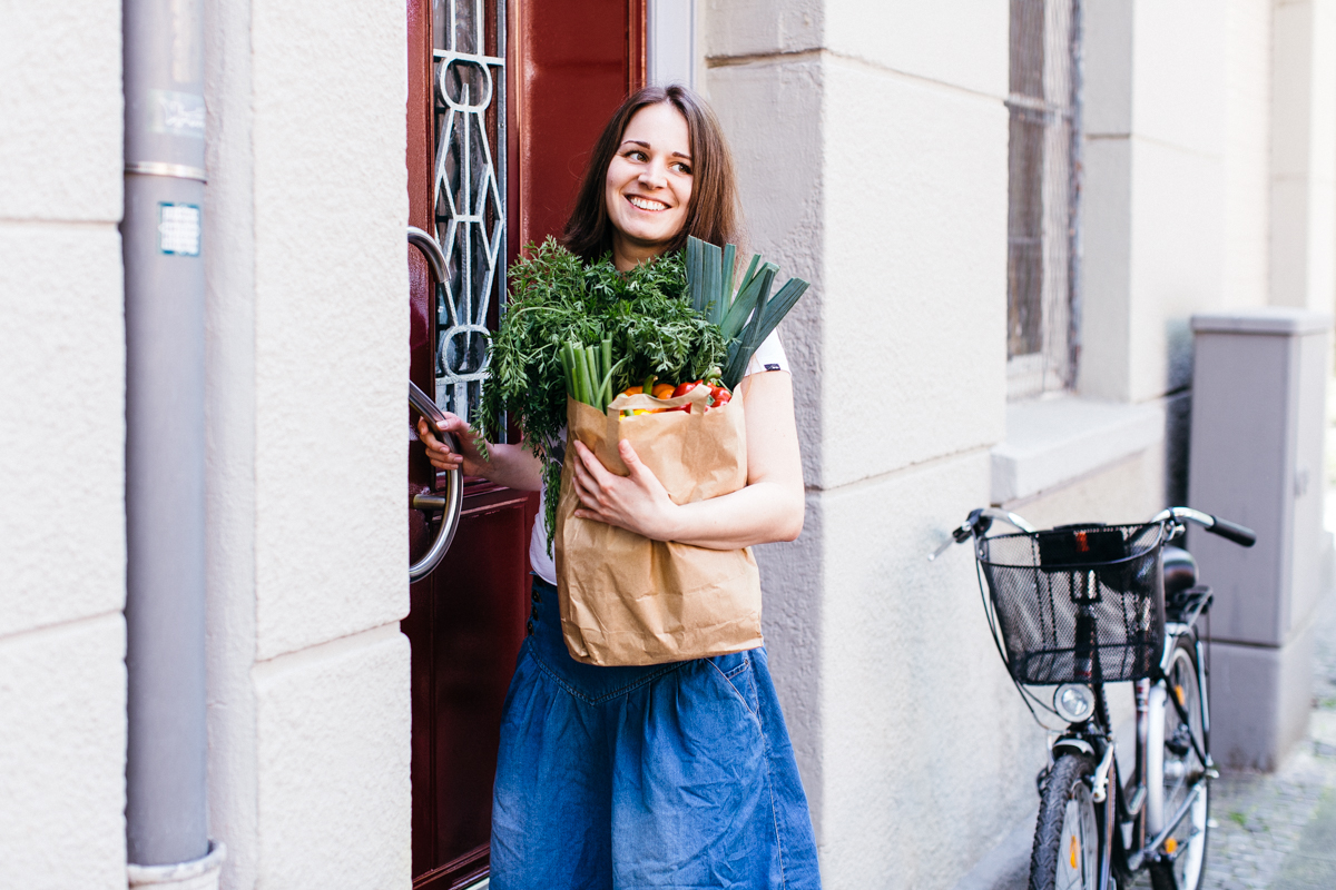 Frau öffnet eine Haustür und trägt eine Papiertüte mit frischem Gemüse und Lebensmitteln, neben ihr steht ein Fahrrad auf dem Gehweg.